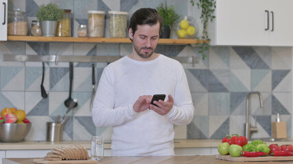 Young Man using Smartphone while Standing in Kitchen