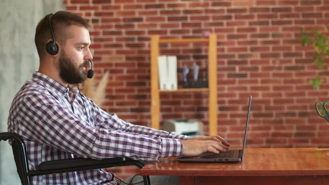 Hipster man in wheelchair use laptop. Disabled person in headset is typing on laptop keyboard, side view. Concept of remote work for people with disabilities