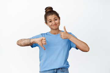 Unsure girl shows thumbs up and down, looking uncertain, like or dislike, standing against white...