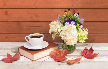 Coffee, book, autumnal white hydrangea, pansy flowers and autumn maple leaves on aged wooden table.