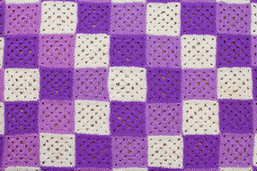Woolen, knitted blanket. Consists of multi-colored squares knitted with woolen threads. The squares are purple, purple, white and sewn together. Textured image for the background. Front view.