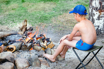 Boy in swimming trunks and cap is warming himself by fire in forest after swimming. Campfire in campsite as means of warming up. Kid warms his frozen feet by fire.