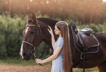 A young beautiful girl with long hair strokes a horse and looks at her in the summer in nature.