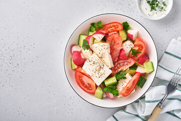 Radish, cucumber, tomato, pepper and feta cheese with spices pepper and olive oil in white bowl on grey slate, stone or concrete background. Healthy food concept. Top view.