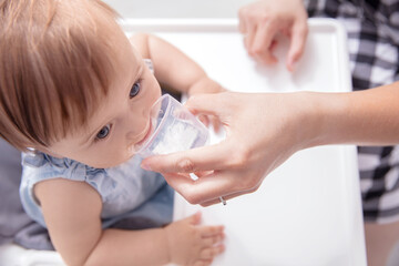 Infant baby girl drinks water from baby cup holded by her mother. View from the top