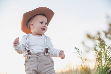 baby in a hat and suspenders looking in the field at sunset