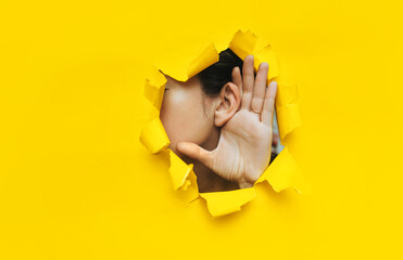 Close-up of a woman's ear and hand through a torn hole in the paper. Yellow background, copy space. The concept of eavesdropping, espionage, gossip and tabloids. - obrazy, fototapety, plakaty