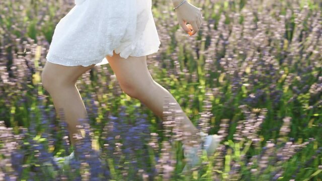 Unrecognizable girl in a white dress running through lavender field. Unknown girl legs running at sunrise meadow. Woman enjoys the fresh air in the countryside. Girl runs on the lavender flowers