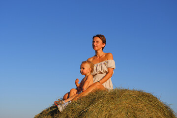 Naklejka na ściany i meble Little boy with mom on a haystack. Mom with baby on a background of blue sky. Mom with a small child on a pile of hay