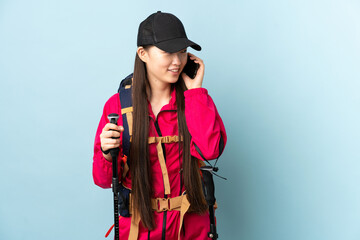 Young Chinese girl with backpack and trekking poles over isolated blue background keeping a conversation with the mobile phone with someone