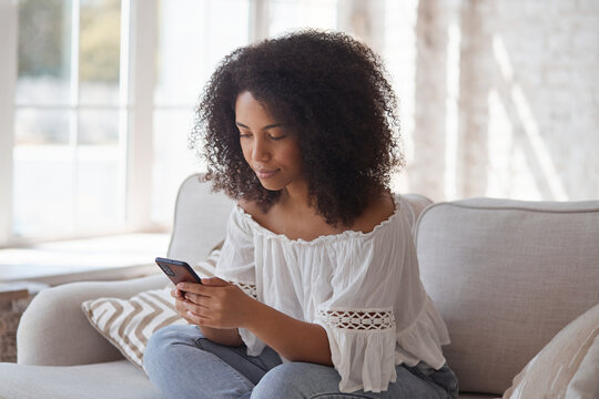 Smiling afro american woman enjoying using smartphone application reading positive news, addiction to mobile phone concept, playing mobile game. shopping and purchasing in mobile delivery app at home