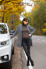 Stylish pretty caucasian woman in glasses, grey coat, brown  sweater and black jeans stands leaning on the SUV car. Autumn forest with yellow leaves on background. Young female travelling on the car.