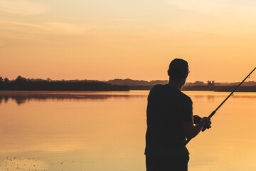 Fisherman recreation with spinning fishing rod outdoor. Handle rotation with reel of fishing rod. Fishing fish in early morning, silhouette on sunrise. Angler stands in river and catching fish