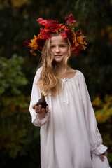 Ein Mädchen mit dem bunten Herbstkranz aus den bunten Blättern auf dem Kopf , blondesteht mit weißem Kleid im Wald. Sie sieht aus wie eine Herbstfee aus. Sie hält ein Frosch mit der Krone in der Hand.