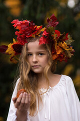 Ein Mädchen mit dem bunten Herbstkranz aus den bunten Blättern auf dem Kopf steht mit weißem Kleid im Wald. Sie sieht aus wie eine Herbstfee aus. Sehr hübsch mit langen, blonden Haaren.