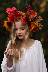 Ein Mädchen mit dem bunten Herbstkranz aus den bunten Blättern auf dem Kopf steht mit weißem Kleid, langen, blonde Haaren im Wald. Sie sieht aus wie eine Herbstfee aus,  In der Hand ist eine Kastanie.
