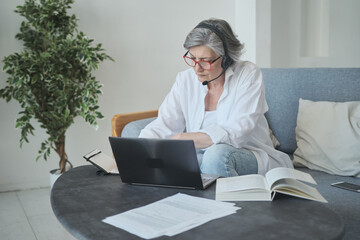 Elderly mature old focused woman wearing headset, studying with teacher online, learning foreign language, making notes in notebook. Internet distant education from home is available at older age