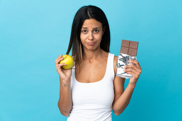 Caucasian girl isolated on blue background taking a chocolate tablet in one hand and an apple in...