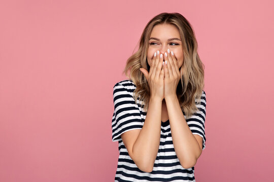 Surprised young woman excited cover open mouth and look side happy laughing over pink studio wall. Young blonde female amazed with happiness in positive shock cheering to good amazing news isolated