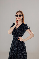 A dark-haired girl in a black dress stands on a white background and straightens her hair with her hand.