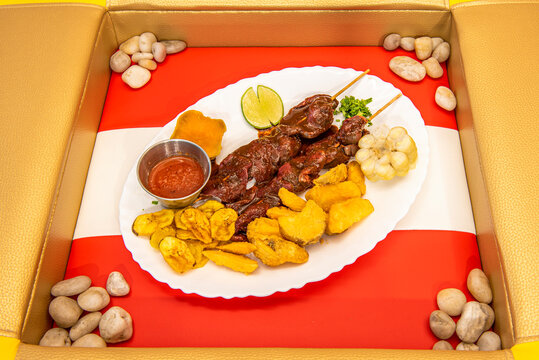 White tray on the flag of Peru with plantain in chips, fried potatoes and potatoes accompanying two pinchos of Peruvian anticuchos cooked on the grill