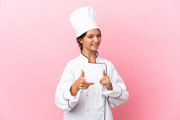 Little caucasian chef girl isolated on pink background pointing to the front and smiling