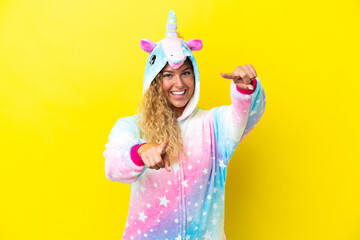 Girl with curly hair wearing a unicorn pajama isolated on yellow background points finger at you...