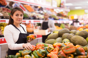 Female supermarket employee in apron lays fresh bell peppers on the counter