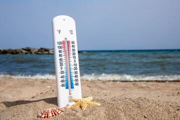 a white thermometer stuck into beach sand with seashells on a hot summer day showing 90 degrees...