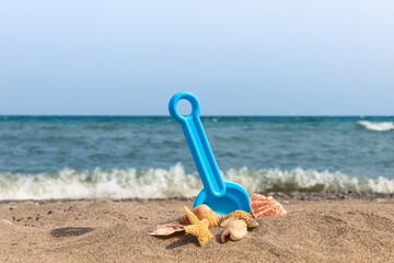 a blue child's toy plastic shovel stuck in beach sand surrounded by seashells and a starfish with a small wave breaking on the beach
