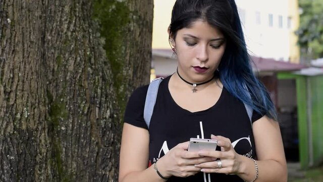 Beautiful serious young woman. blue haired college girl looking at her social media in a park, next to a tree. girl looking at her cell phone in a park. Technology, communication and education concept