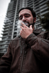 Adult Hispanic man with a beard wearing a leather jacket and smoking a cigarette outdoors