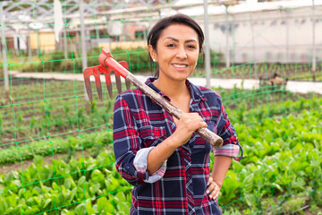Attractive woman gardener worker standing with rake and smiling at greenhouse farm