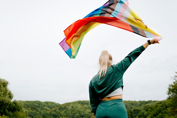 Back view of young woman waving rainbow flag against sky outdoors, new symbol of LGBTQ + community,...