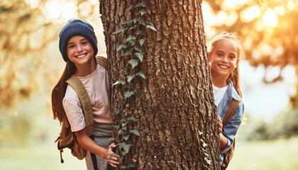 Happy school girls hugging tree in forest and smiling at camera during camping activity in nature
