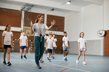 Young female coach has PE class with group of elementary students at school gym. - obrazy, fototapety, plakaty
