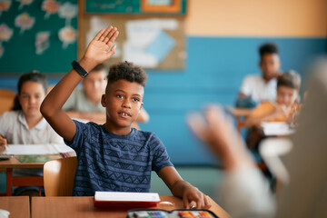 Black elementary student raises his hand to answer question during class at school. - obrazy, fototapety, plakaty