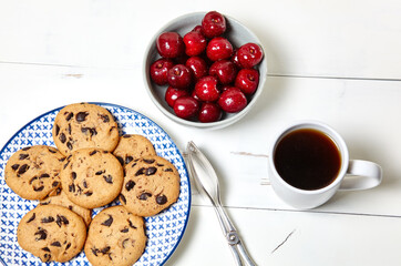 White cup with coffee, traditional cookies on plate and sweet cherries in a ceramic bowl on a white wooden background, closeup. Tasty breakfast, morning routine concept