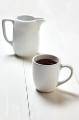 White cup with coffee and milk jug on a white wooden background, closeup. Energy breakfast, morning routine concept