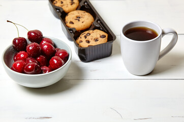 White cup with coffee, traditional cookies and sweet cherries in a ceramic bowl on a white wooden background, closeup. Tasty breakfast, morning routine concept