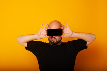 Photo of a joyful bearded hipster holding a smartphone over his eyes. place to advertise the phone, place for text. isolated on yellow background