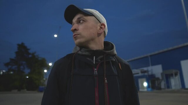 Portrait of a Man in a Baseball Cap Among the Night City Lit by Lanterns. Floored Camera Movement. A Man at Night Looks Around Standing Among Warehouse Buildings