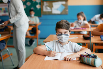 Happy elementary student wears face mask and writes during class at school while looking at camera.