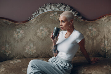 Millenial young woman with short blonde hair portrait sitting with cup of tea english drink on vintage sofa