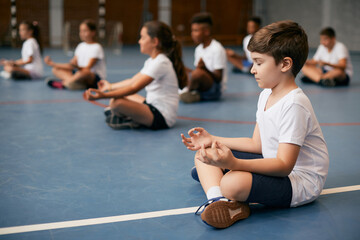 Schoolboy and his friends meditate while practicing Yoga on PE class at school gym.