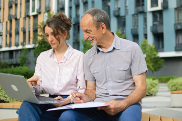 Attractive young woman and senior man are using laptop sitting on the bench in modern residential complex. Casual meeting outside office. A daughter is teaching technology for her father