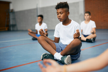 Black elementary student does breathing exercise during Yoga class at school gym. - obrazy, fototapety, plakaty
