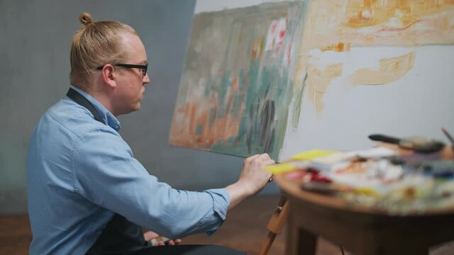 Artist's workshop, creative person at work, a talented adult man artist covers the canvas with oil paint, draws the modern abstract picture, 4k slow motion.