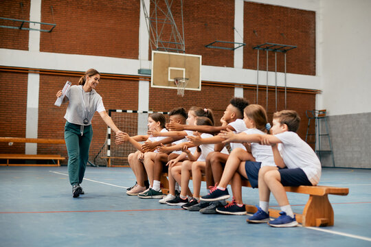 Happy sports teacher greets with group of elementary students during PE class at school gym.