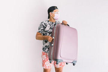 Young man wearing a mask to protect himself from covid-19 on vacation going on a trip, wearing a floral shirt and cap, holding a suitcase, in daylight on a white wall